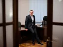 Lex Tarrel в деловом костюме сидит на краю стола в рабочем кабинете, 2016 год, вид через дверь